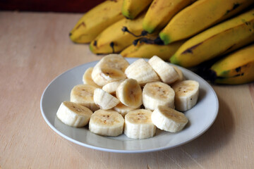 Banana slices on a white dish and bananas fruit in the background.