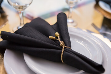 Detalhe de guardanapo de tecido preto com enfeite dourado, dentro do prato.