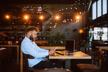 A young handsome caucasian businessman with a beard is sitting in a cafe drinking whiskey and working on a laptop while using the phone. Work in a restaurant