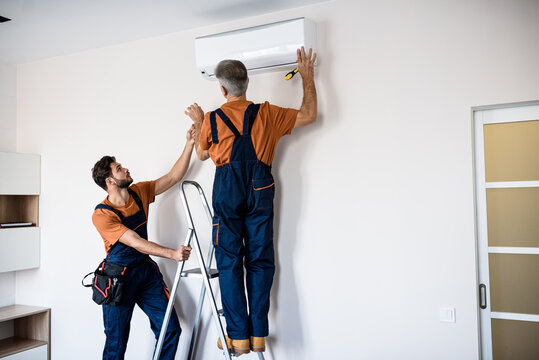 Two workers in uniform, air conditioning masters using ladder while installing a new air conditioner in the apartment. Construction, maintenance and repair concept