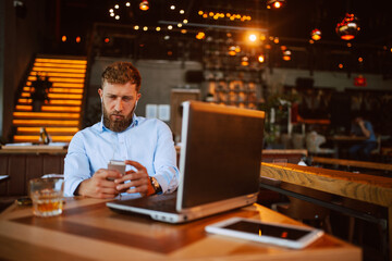 A young handsome caucasian businessman with a beard is sitting in a cafe drinking whiskey and working on a laptop while using the phone. Work in a restaurant