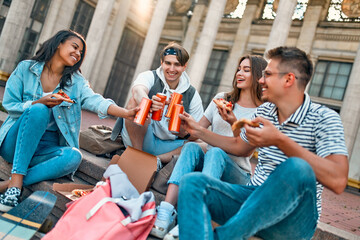 A group of students sit on the steps outside the campus and eat pizza and soda. A group of friends are relaxing and chatting.