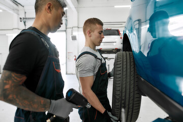 Giving your car a best treat. Male mechanic holding car wheel, while his colleague screwing or unscrewing wheels by pneumatic wrench at auto repair shop