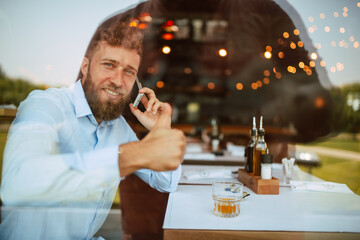 A young handsome caucasian  businessman with a beard is sitting in a cafe drinking whiskey and talking on the phone. Work in a restaurant