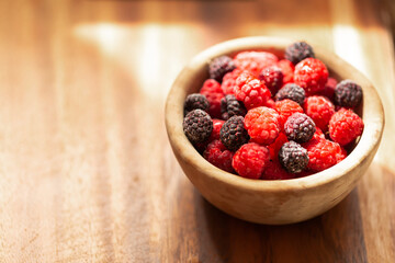 Red and black raspberries on a wooden plate. Ripe and fresh organic berries on a wooden background. Sunlight falls from the window. Blurred background.
