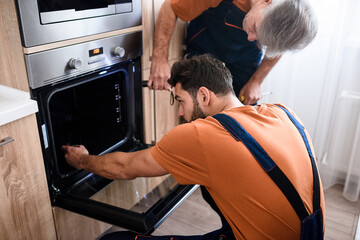 Close up shot of two repairmen, workers in uniform examining oven using screwdriver and flashlight indoors. Repair service concept