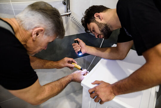 Close up of two repairmen, workers in uniform working, fixing washing mashine using screwdriver and flashlight in the bathroom. Repair service concept