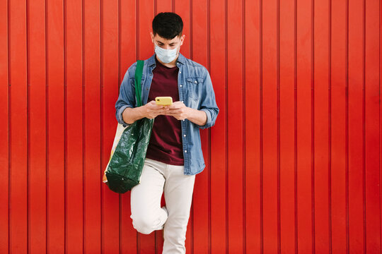 Man with protective mask and shopping bag looking at cell phone in front of red background