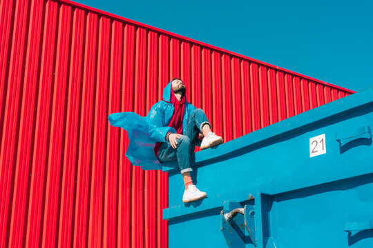 Young man wearing rain coat sitting on edge of blue container in front of red wall