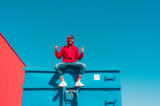 Young man wearing red hoodie, sitting on edge of blue container, making sign of the horns