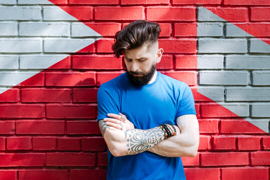Fototapeta Young man with tattoo standing in front of red brick wall with arms crossed