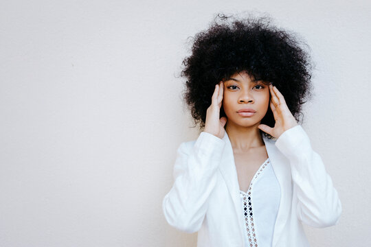 Black womam, wearing white suit, standing in front of white wall, with hands on forehead