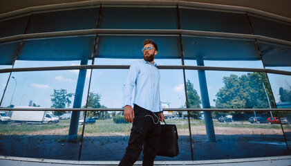 A young caucasian businessman with sunglasses is standing in front of the airport with a black briefcase in his hand