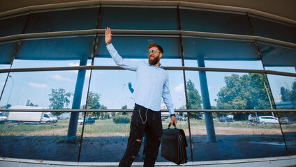 A young caucasian  businessman with sunglasses stands in front of the airport with a briefcase in his hand and waves