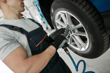 Cropped shot of male mechanic screwing or unscrewing car wheel of lifted automobile by pneumatic...
