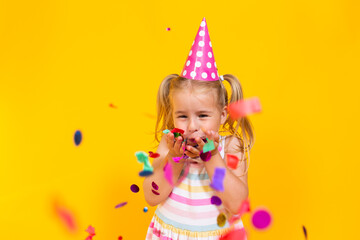 Happy birthday child girl in pink cup blowing confetti on colored yellow background. Celebration, childhood.
