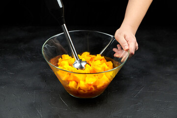 Pumpkin pieces and blender in a glass bowl on black table. Chopped butternut squash, cooking vegetable food, pumpkin puree (mash) recipe