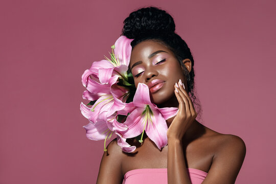 Art make up. Fashion beauty portrait of young African American model touching face with posing with lily flowers against pink background.