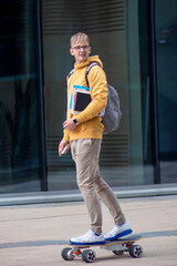 Handsome guy, young man, hipster, student or pupil in glasses on his face riding on electric urban modern skateboard with backpack, books and textbooks. Eco transport, university, technology concept. © Евгений Шемякин