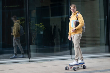 Handsome guy, young man, hipster, student or pupil in glasses on his face riding on electric urban modern skateboard with backpack, books and textbooks. Eco transport, technology concept. © Евгений Шемякин
