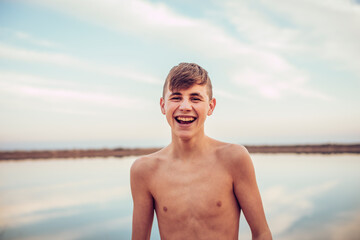 Portrait of happy shirtless teenage boy standing with lake and cloudy sky in background