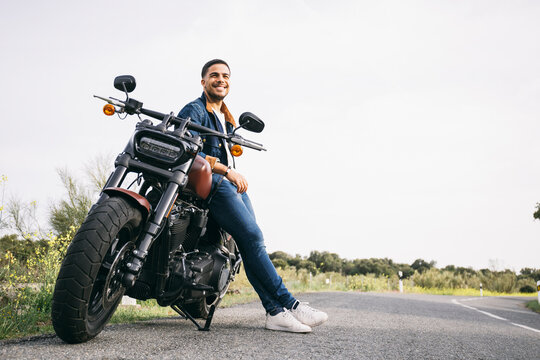 Smiling young biker leaning on motorcycle on road against sky