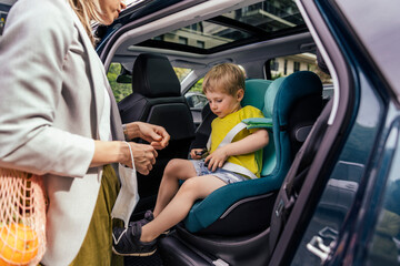 Little boy fastening seat bell of child's seat in car while his mother watching him