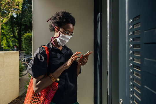Young woman wearing textile protective mask standing in front of entry door using smartphone