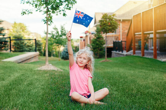 Adorable cute happy Caucasian girl holding Australian flag. Smiling child sitting on grass in park holding Australia flag. Kid citizen celebrating Australia Day holiday in January outdoors.