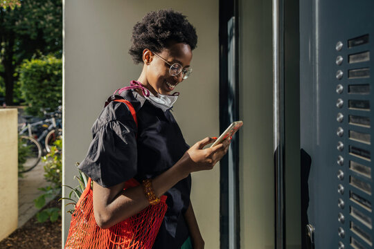 Smiling young woman with textile protective mask standing in front of entry door looking at smartphone
