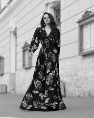 Young beautiful lady wearing black dress with colorful floral design and standing at city street. Pretty woman outdoor full length portrait. Fashion model.