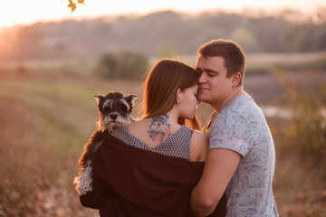 A couple in love hugs, kisses in the rays of the autumn sun, with their backs turned to the camera, holding a Schnauzer in their arms. A beautiful girl has a dog portrait tattoo on her back. Family