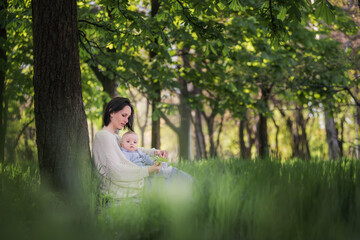 Modern beautiful young brunette mother with a short haircut, holding a toddler in a gray knitted romper in her arms. A girl and her son are sitting under a tree, playing together, hugging. Mom kisses 