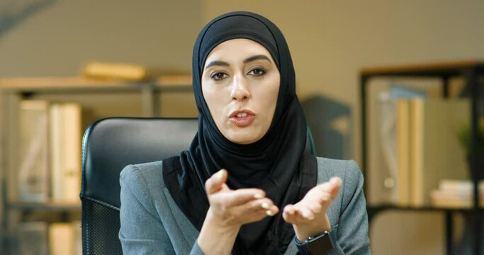 Young beautiful muslim woman in black hijab sitting at table in office and talking to camera while educating. Charming pretty Arabian business blogger or cauch in traditional headscarf speaking.