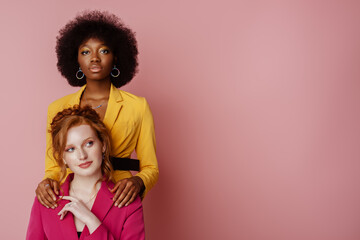 Diverse beauty, fashion: portrait of two multiethnic women wearing colorful blazers, posing on pink...