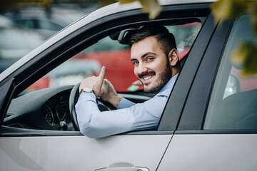 Handsome man sitting in luxury car in front of a steering wheel smiling and pointing up