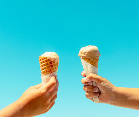Couple kids hands holding ice cream cones against blue sky background in summer vacation. Summer food, summertime joy, vacation, holidays.