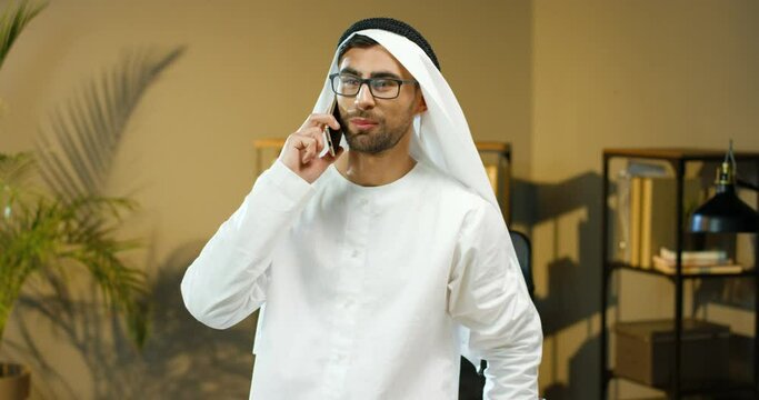 Young joyful Arabic man in Kandura and glasses standing in office in UAE, talking on mobile phone and smiling. Male muslim working and speaking on cellphone in cabinet. Arab businessman in dish dasha.