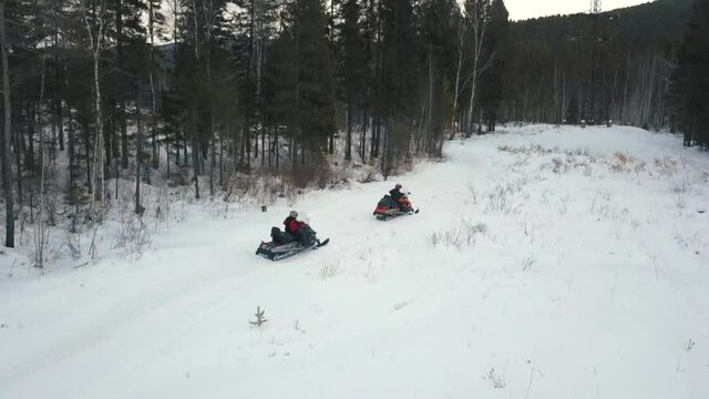 Aerial view of row of snow mobiles ride on a snowy trail near the forest. Clip. Concept of winter extreme sport, people riding snow mobiles along the trees under the heavy sky.