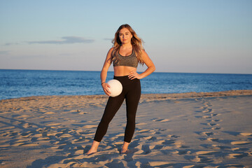 Stunning young blonde woman with volleyball on beach at sunset
