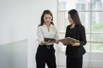 Two female office workers in business suit discuss about their work with tablet and file in hand in business building. Businesswoman talking and poitning on tablet. Business stock photo