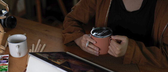 Female artist holding coffee cup while working with tablet on worktable with painting tools