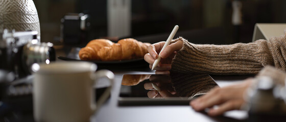 Female working with digital tablet on worktable with camera, supplies, coffee cup and croissants