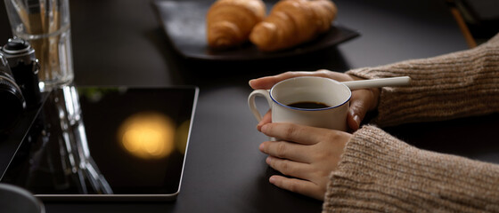 Female holding coffee cup on dark office desk with digital tablet, croissant, camera and supplies