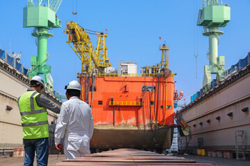 Worker in shipyard for ship repair in floating dry dock yard for recondition maintenance with sand blasting perform, port control and inspector surveying condition in background - obrazy, fototapety, plakaty