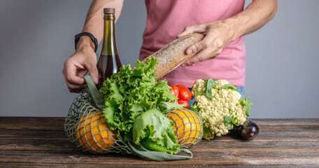 Modern young man is pulling out purchases from a string bag on a wooden table. Concept of eco-friendly behavior, the use of reusable bags and concern for the environment