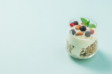 A large glass with granola, yogurt and berries on a light blue background.