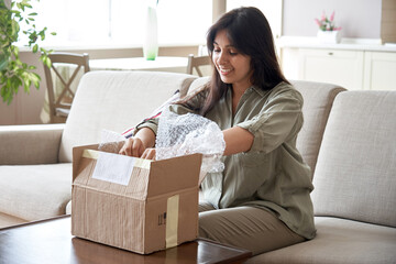 Smiling indian woman shopper customer opening post package box sitting on couch at home. Happy...