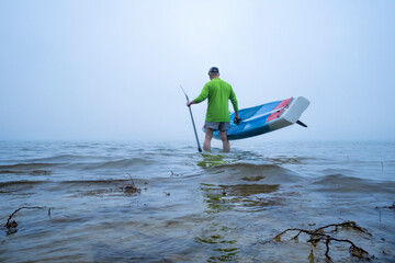 Naklejka premium mature male paddler is launching stand up paddleboard on a foggy lake - Boyd Lake State Park in northern Colorado, a popular boating and recreation destination.