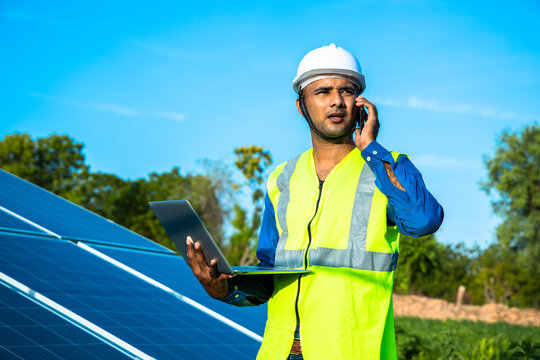 Young Male Technician Worker Wearing Green Vests And Helmet With Laptop In Hand Talking On Phone About The Maintenance And Installation Of The Solar Panels Standing In Field, Technology In Agriculture